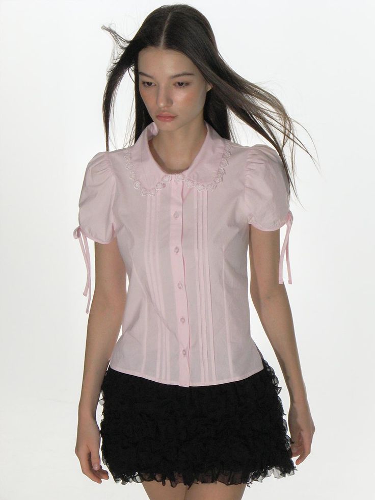 This product is a finely crafted blouse with a lace collar that exudes a vintage charm. It features pintuck detailing along the front, adding texture and a delicate femininity to the overall design. The short sleeves with tie details offer a customizable fit and contribute to the blouse's romantic and timeless appeal. - This blouse is adorned with a lace collar that frames the neckline beautifully, creating a subtle statement.- Pintuck details provide a structured yet soft appearance, enhancing the garment's refined look.- The short sleeves finished with ties allow for a bow accent, lending an air of whimsy to this classic piece.- Constructed with a button front closure, it offers ease of wear while maintaining a polished aesthetic. Pink Lace Trim Blouse, Elegant Pink Short Sleeve Tops, Feminine Blouse With Collared Neckline, Fitted Feminine Top With Doll Collar, Fitted Pink Blouse With Lace Collar, Feminine Formal Top With Peter Pan Collar, Formal Feminine Top With Peter Pan Collar, Feminine Formal Tops With Peter Pan Collar, Chic Pink Top With Lace Collar