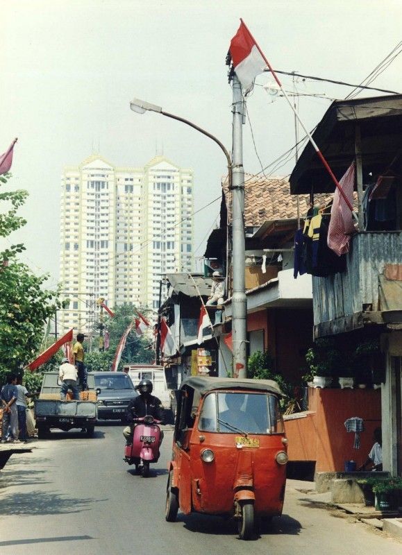 an old red car driving down a street next to tall buildings with flags on them