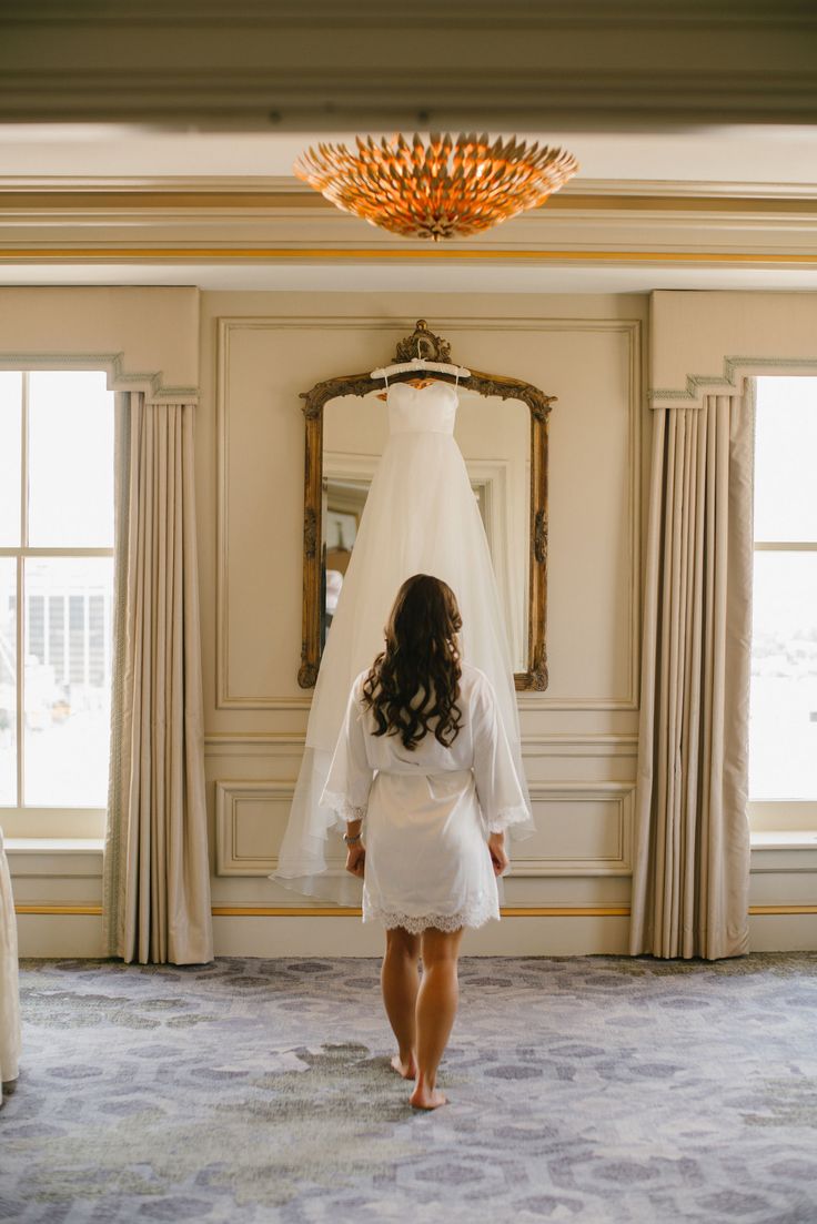 a woman standing in front of a wedding dress hanging on a wall next to a window