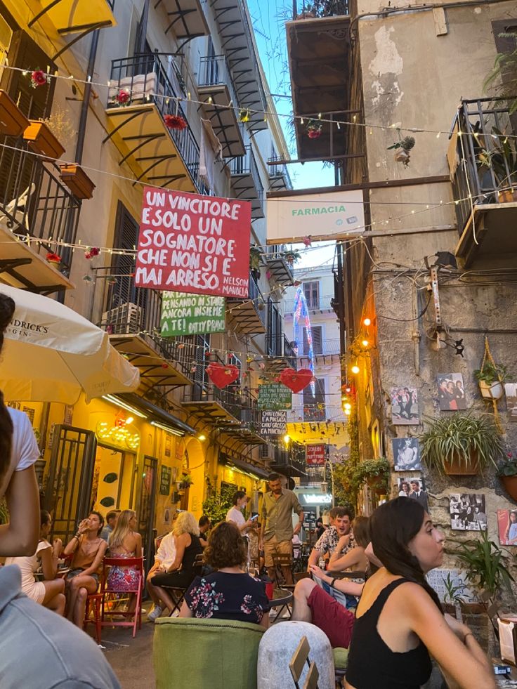 people sitting at tables in an alleyway with signs hanging from the buildings above them