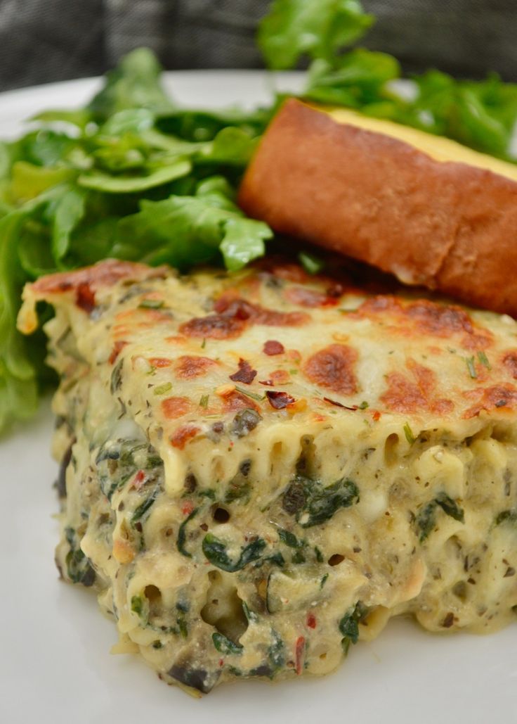 a white plate topped with lasagna covered in cheese and spinach next to a hot dog bun