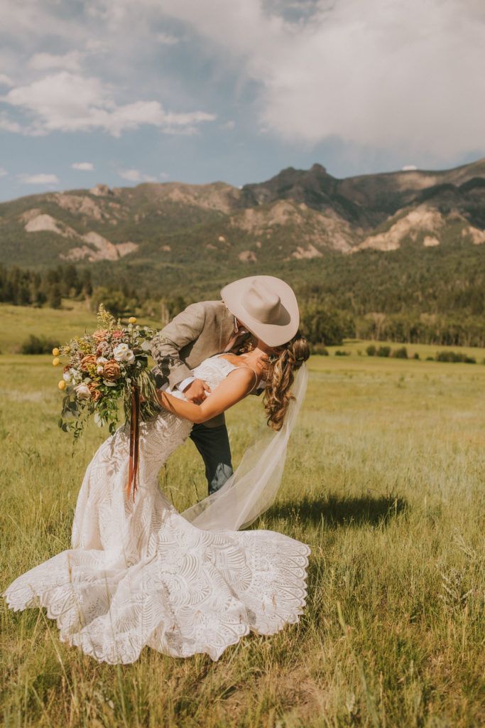 a bride and groom kissing in a field with mountains in the background at their wedding