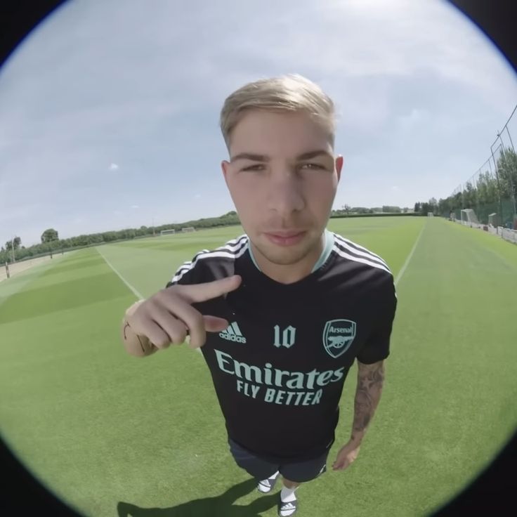 a young man is standing in front of a soccer field and pointing at the camera