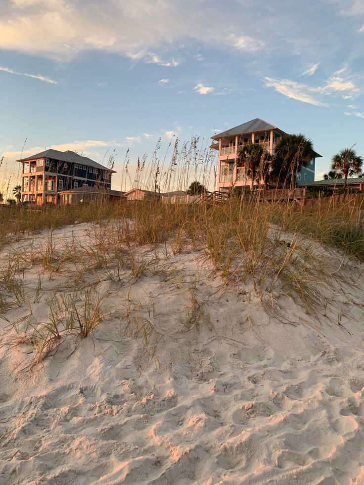 two beach houses are in the distance behind some tall grass and sand on a sunny day