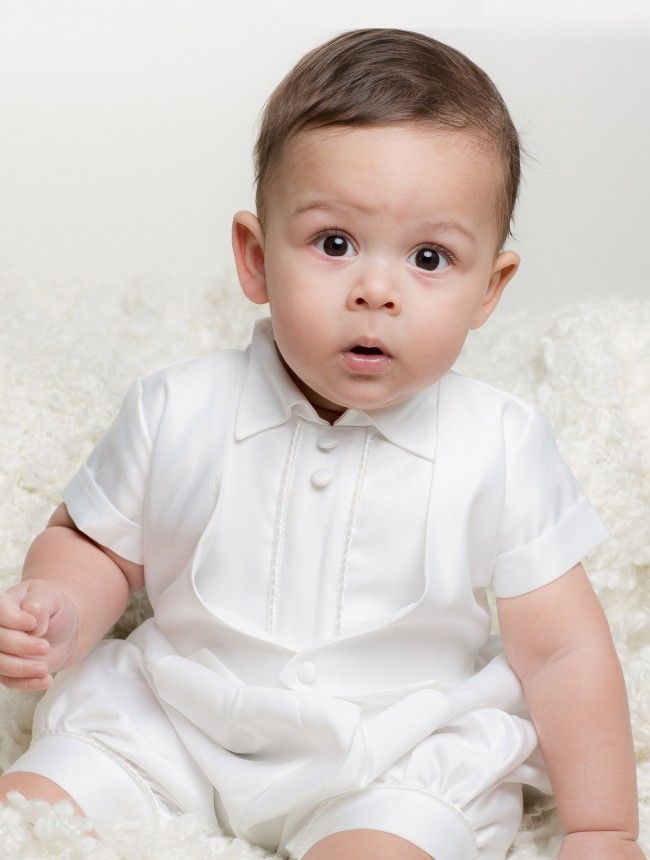 a baby in a white shirt and dress sitting on a bed looking at the camera