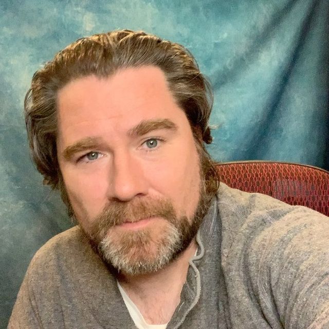 a man with long hair and beard sitting in front of a blue background wearing a gray shirt