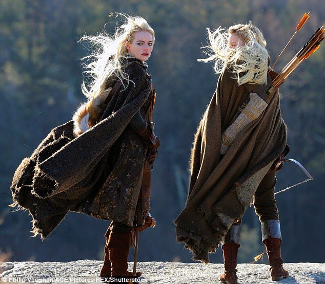 two women dressed in medieval clothing holding arrows and standing on a hill overlooking the woods