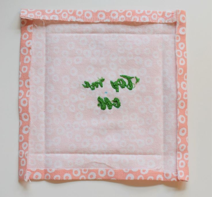 a pink and white square with embroidered words on the front, two small green leaves in the middle