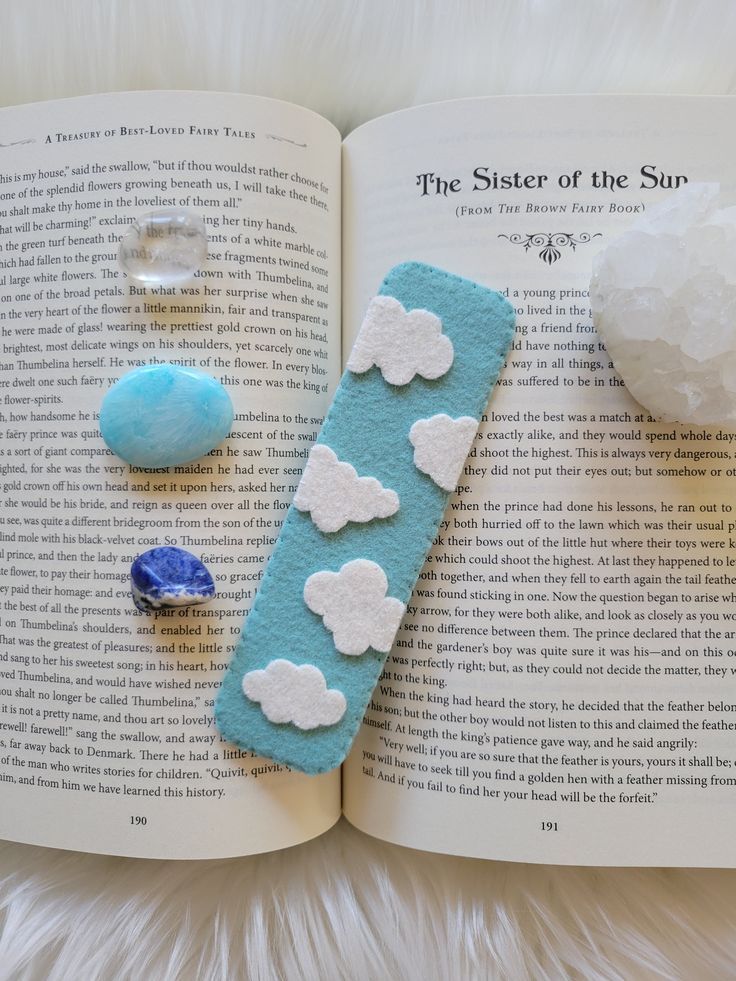 A light blue bookmark with white clouds sitting ontop of a book, surrounded by crystals. Gifts With Wool, Handmade Felt Bookmark, Easy Felt Patterns, Book Marks Felt, Bookmark Clouds, Kids Bookmark Craft, Small Felt Projects, Diy Felt Bookmarks, Felt Bookmarks Diy Pattern