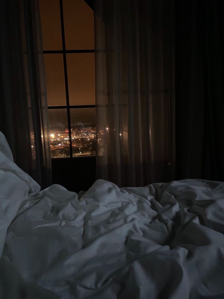 an unmade bed with white sheets and city lights in the distance from a window