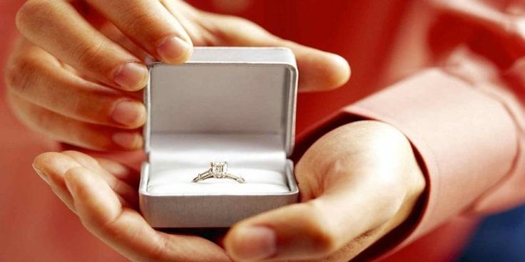 a person holding an engagement ring in their left hand and wearing a red shirt behind them