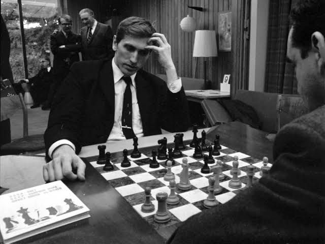 a man sitting at a table playing chess with another man in the background looking on