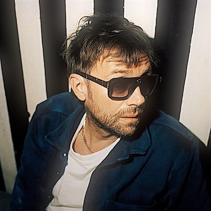 a man wearing sunglasses sitting in front of a black and white striped wall with his head tilted to the side