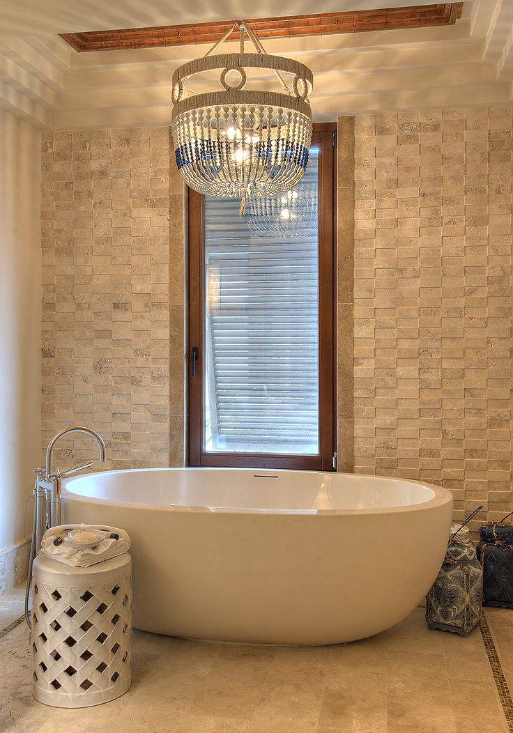 a white bath tub sitting under a chandelier next to a window in a bathroom