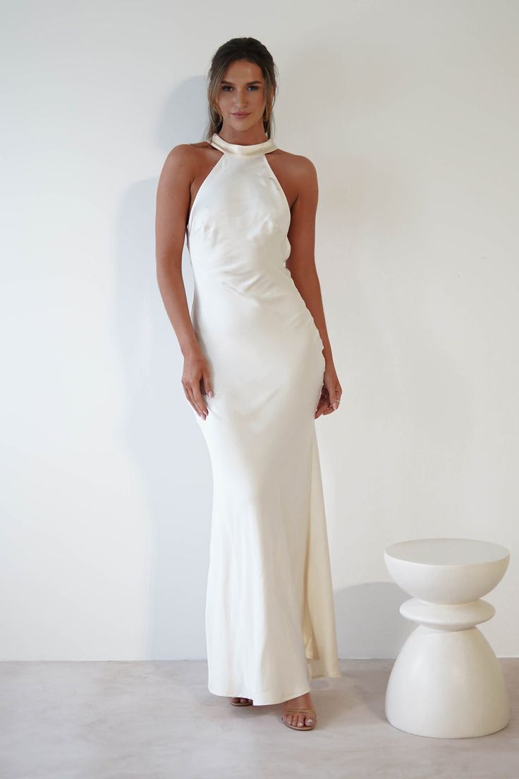 a woman standing in front of a white wall wearing a long dress with halter neck