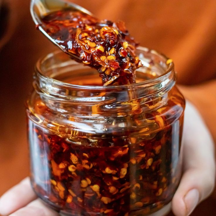 a person is holding a spoon full of food in a glass jar with some liquid on it