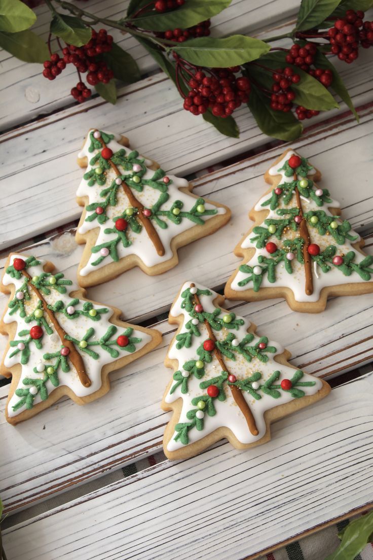 four decorated christmas tree cookies sitting on top of a white wooden table next to holly wreaths