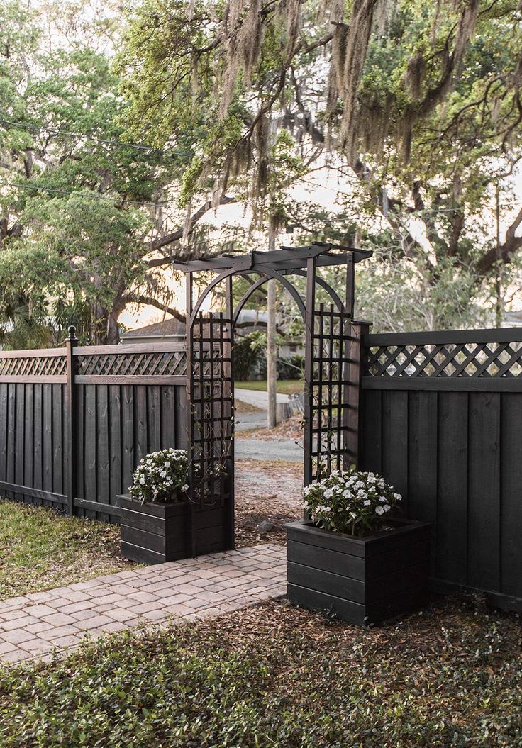 two black planters with white flowers are in front of a wooden fence and gate