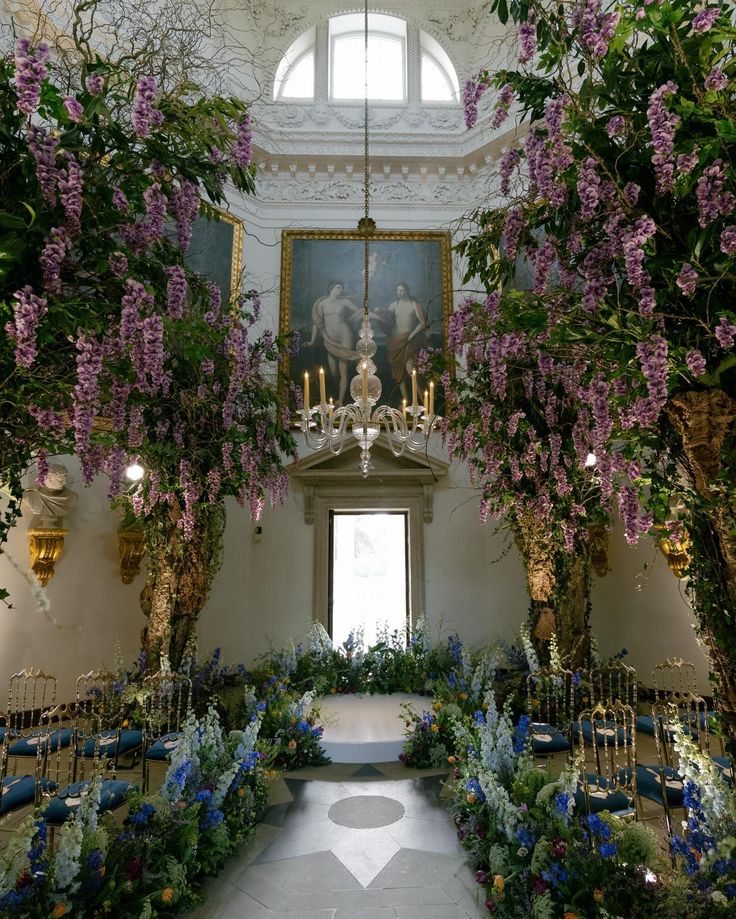 an indoor ceremony with purple flowers and greenery on the ceiling, chandelier hanging from the ceiling