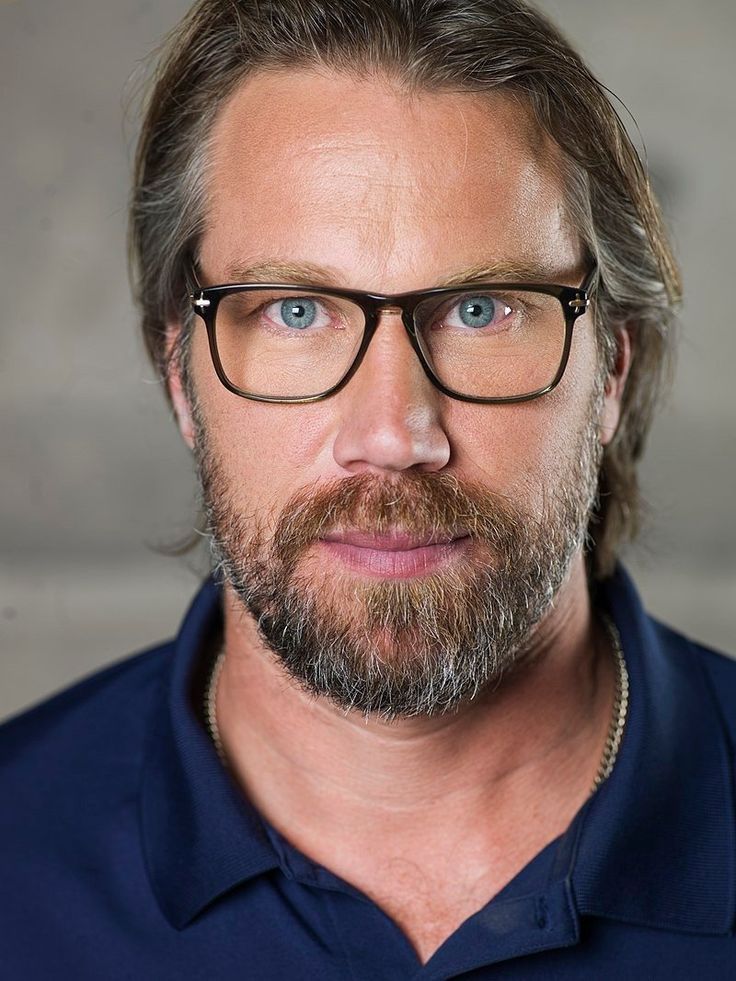 a man with long hair and glasses looks at the camera while wearing a blue shirt