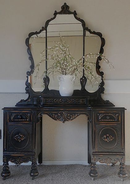 an ornate black desk with mirror and vase on top, in front of a wall