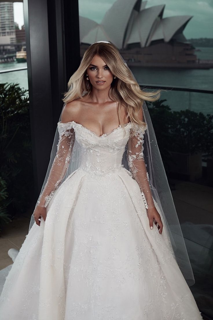 a woman in a white wedding dress standing next to a window with the sydney harbour bridge in the background