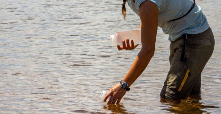 a woman is standing in the water holding a plastic cup and looking at something on her hand