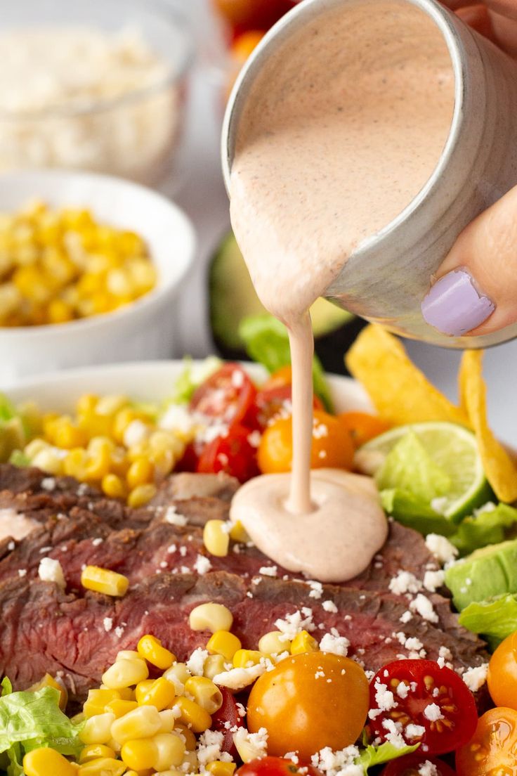 a person pouring dressing onto a steak salad with corn, tomatoes and lettuce