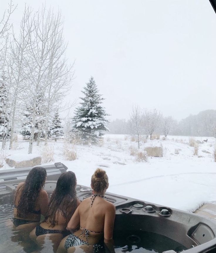 three women sitting in a hot tub with snow on the ground and trees behind them