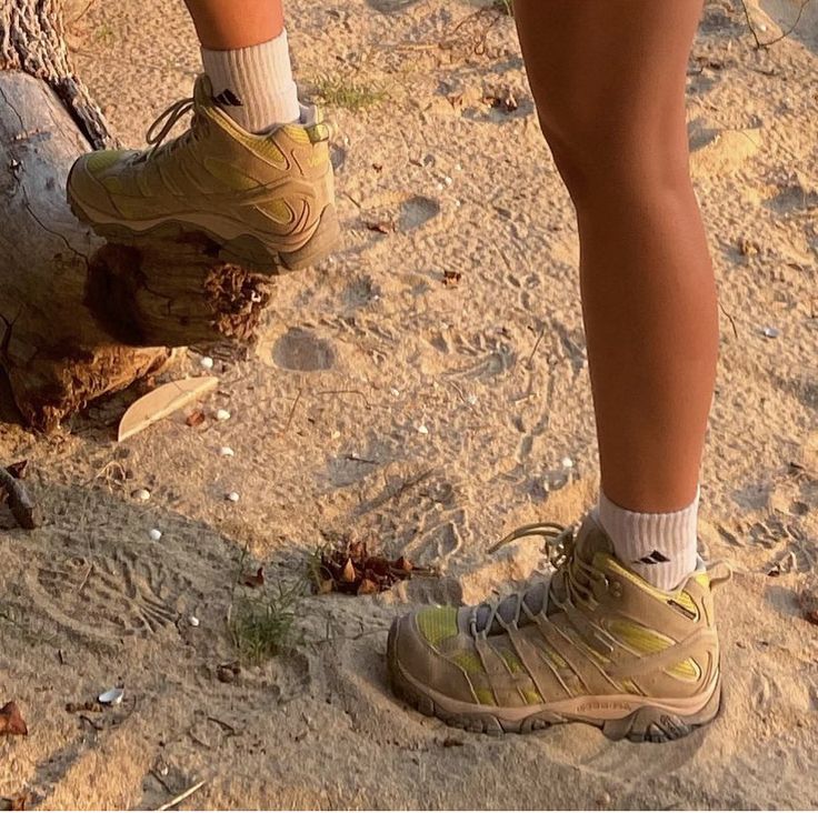 a person standing on top of a sandy beach next to a tree trunk and wearing hiking shoes