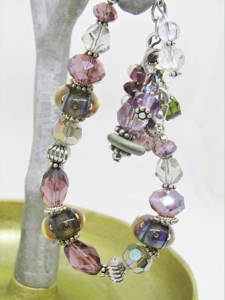 "This ARTISAN GLASS (Lampwork) BRACELET features 3 BOROSILICATE GLASS BEADS in shades of purple and green. The Boro glass beads are roughly 8x15mm and are spaced throughout the bracelet. There are also 10x14mm crystal tapered faceted ovals in purple and 8x12mm faceted crystal rondelles in the same purple; 5x8mm silver spacer beads, along with assorted clear and lavender faceted crystals. There are 7 assorted drops on the chain, inclding lampwork rondelles, Swarovski crystals, other glass, crysta Adjustable Hand-strung Beaded Bracelets With Czech Glass, Hand-strung Czech Glass Crystal Bracelet As Gift, Bohemian Purple Czech Glass Bracelets, Useable Gifts, Artisan Czech Glass Hand-strung Beaded Bracelets, Adjustable Czech Glass Crystal Bracelet, Hand-strung, Lampwork Bracelets, Farmers Market Bag, Green Bracelet