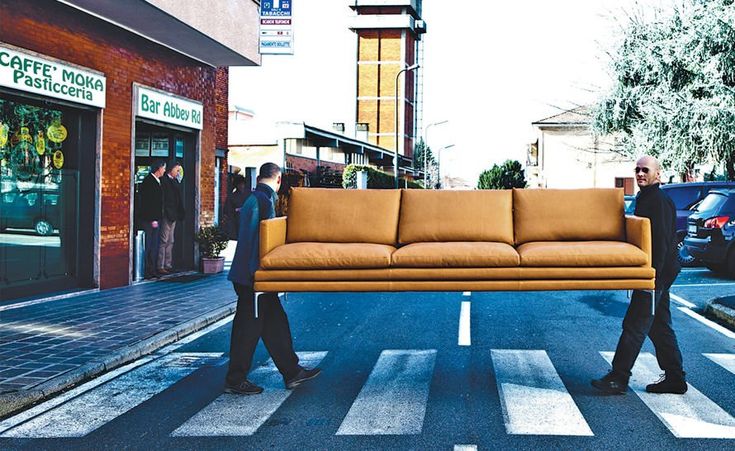 two men walking down the street carrying a couch across the street in front of them