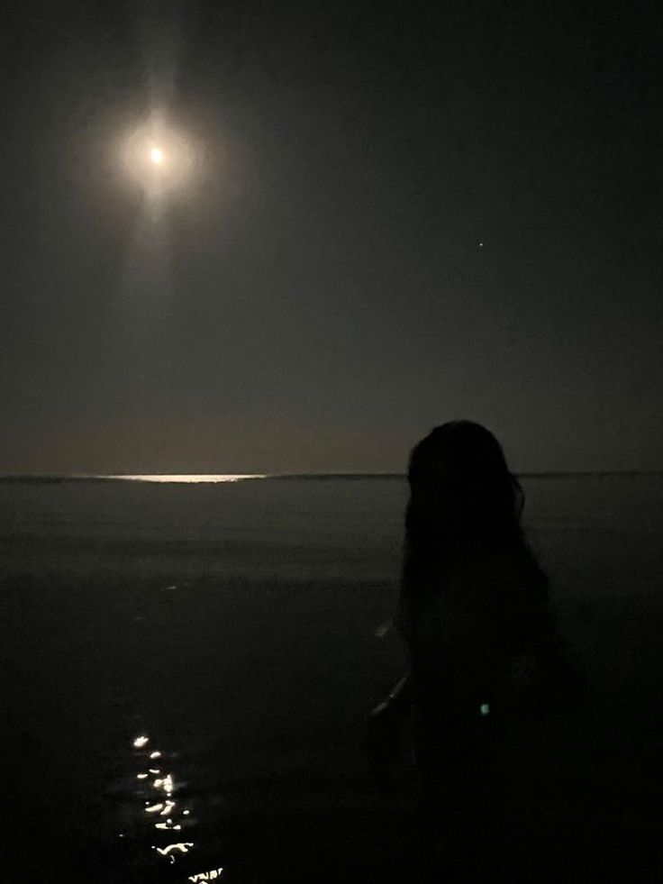 a person standing in the water at night with the moon shining over the ocean behind them