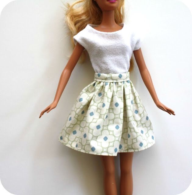 a doll with blonde hair wearing a white top and blue polka dot skirt, standing next to a wall