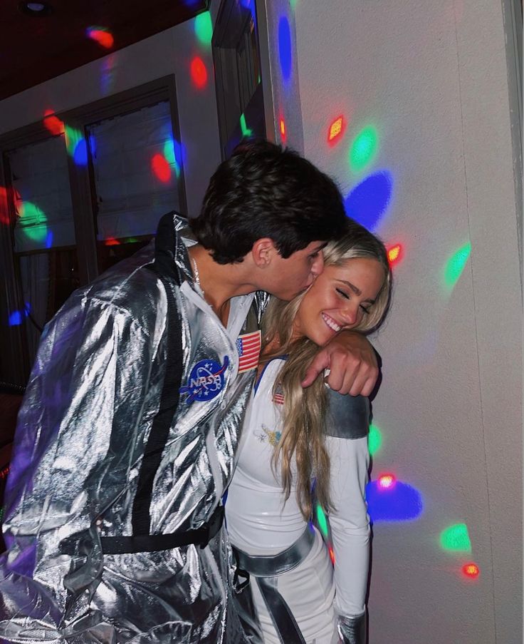 a man and woman dressed in silver posing for a photo with colorful lights on the wall behind them