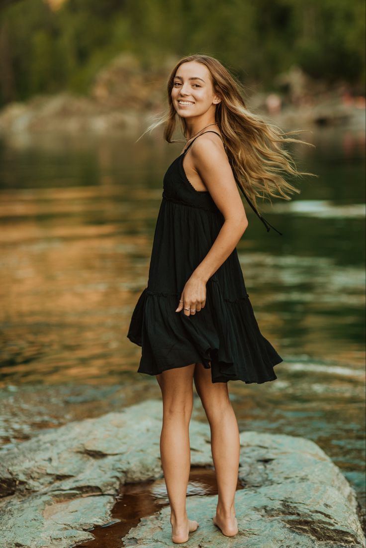 a beautiful young woman standing on top of a rock next to the water with her hair blowing in the wind