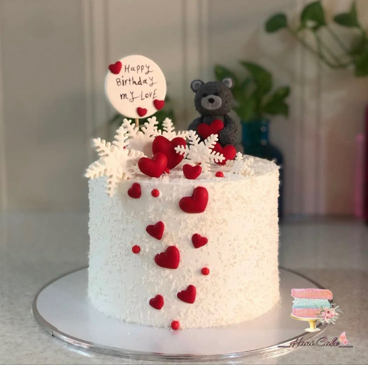 a white cake with frosting and red hearts on it, topped with a teddy bear