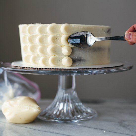 a person is holding a spoon over a cake on a glass platter with white frosting