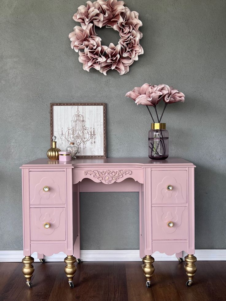 a pink desk with two drawers and flowers on it in front of a gray wall