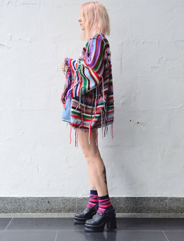 a woman standing in front of a white wall wearing black boots and a colorful sweater