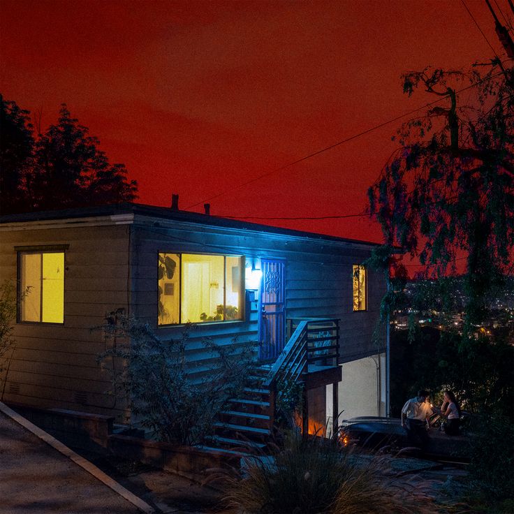 two people standing in the window of a small house at night with red sky behind them