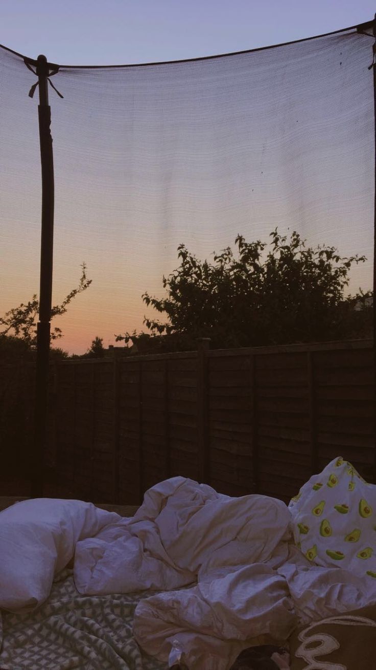 an unmade bed in front of a fence at sunset