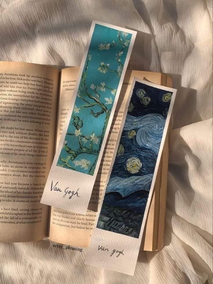 two bookmarks with the same painting on them are laying next to an open book