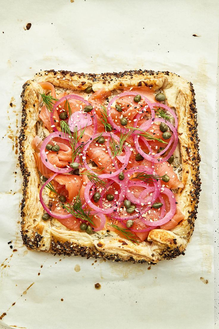 a square pizza with onions, tomatoes and herbs on it sitting on a piece of parchment paper