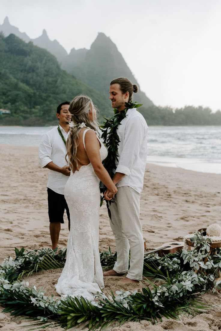 a man and woman standing on top of a sandy beach next to the ocean with greenery