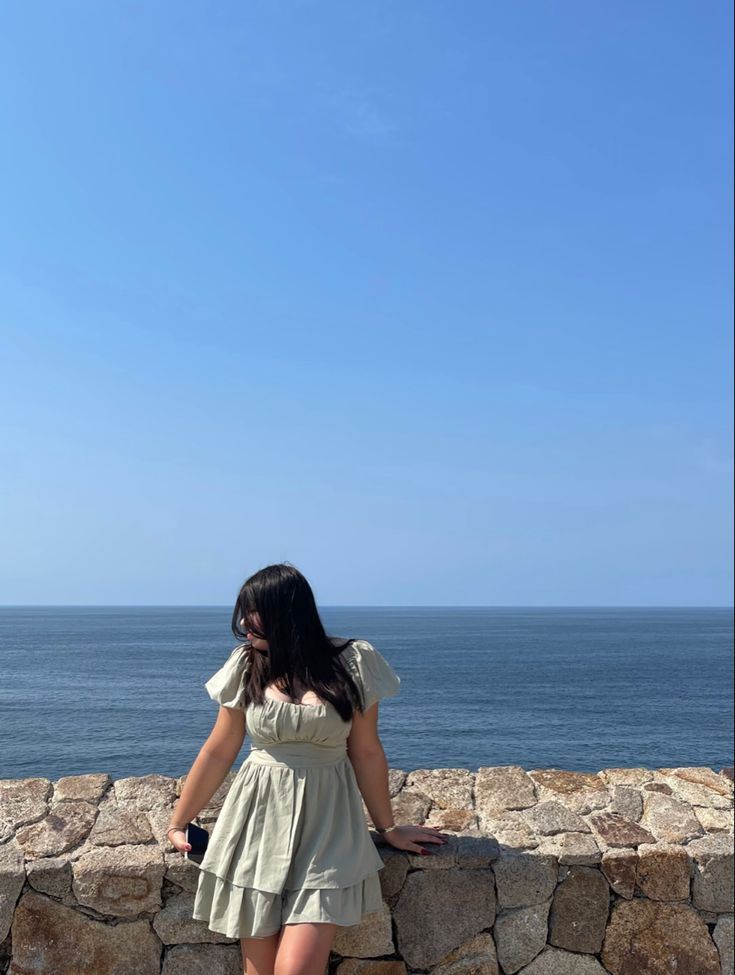 a woman standing on the edge of a stone wall looking at the ocean in front of her