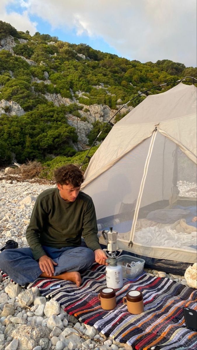 a man sitting in front of a tent on top of a rocky beach next to a picnic table