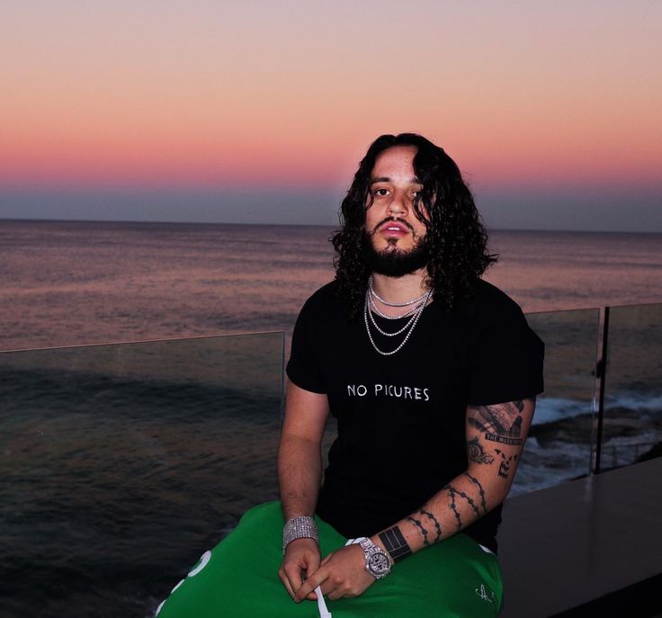 a man with long hair sitting on top of a green pillow next to the ocean