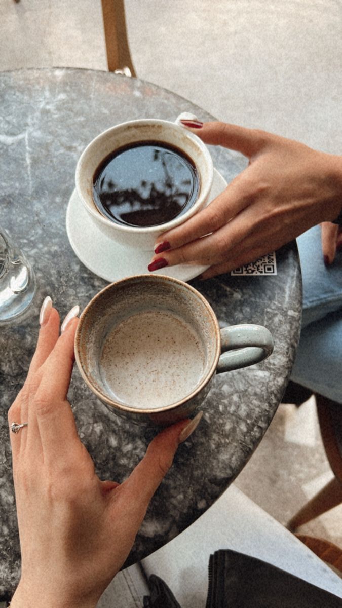 two people sitting at a table with cups of coffee in front of them and their hands on the cup