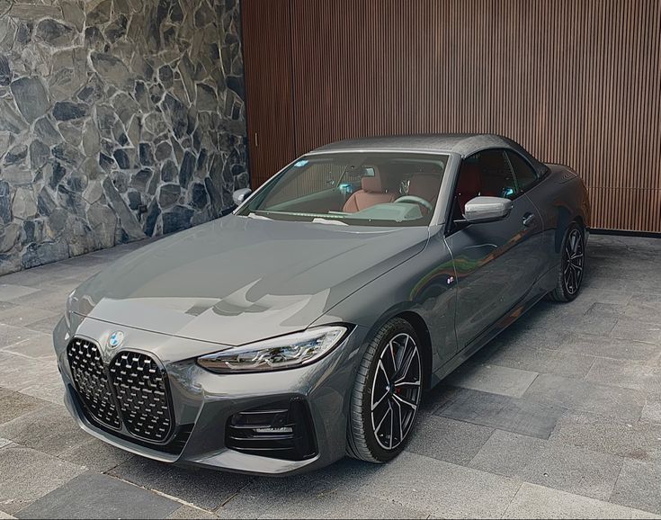 a grey bmw car parked in front of a stone wall with two people looking out the window